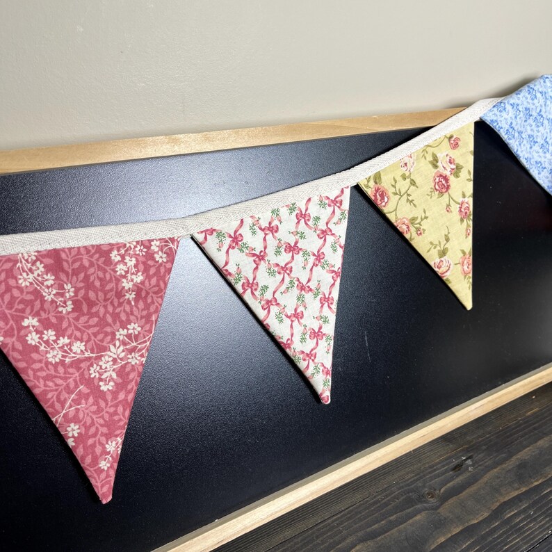 May include: A decorative banner with triangular fabric flags. The flags feature floral and bow patterns in shades of pink, yellow, and blue, strung along a natural-colored ribbon. The banner is displayed against a black background.