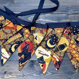 May include: A Halloween-themed fabric banner with triangular pennants. Each pennant features a unique design with pumpkins, witches, moons, and the text "Happy Halloween". The banner has a dark blue trim and a matching ribbon for hanging.