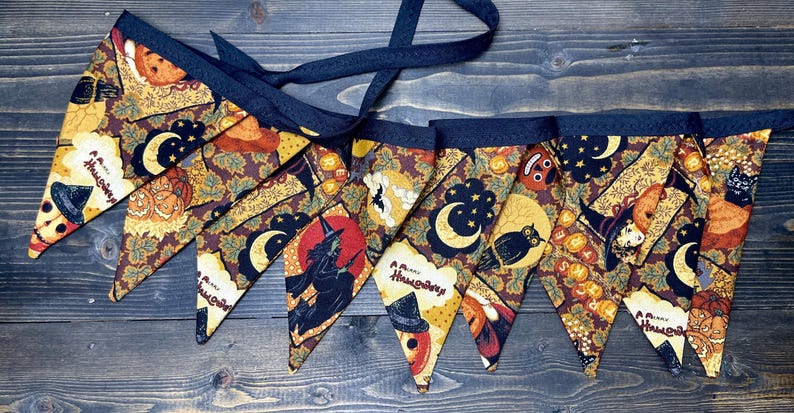 May include: A Halloween-themed fabric banner with triangular pennants. Each pennant features a unique design with pumpkins, witches, moons, and the text "Happy Halloween". The banner has a dark blue trim and a matching ribbon for hanging.