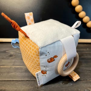 May include: A soft, multi-textured baby toy cube with a white, plush top, a blue and white fabric side with a fish design, a yellow and orange honeycomb fabric side, and a wooden ring attached to a white ribbon. The cube has a tag that reads "The Polka Dot & Spot".