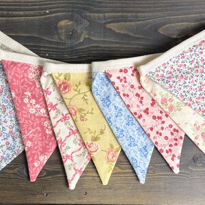 May include: A colorful fabric bunting with various floral patterns in pink, blue, yellow, and white. The pennants are strung together with a natural-colored ribbon, creating a decorative banner for celebrations or home decor.