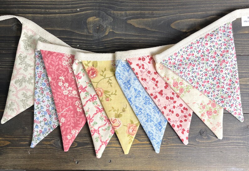 May include: A colorful fabric bunting with various floral patterns in pink, blue, yellow, and white. The pennants are strung together with a natural-colored ribbon, creating a decorative banner for celebrations or home decor.