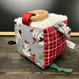 May include: A soft, square baby toy with a wooden ring and red ribbon. The cube features gray fabric with snowmen and red birds, and red plaid fabric. The top is white, fluffy fabric, and a green fabric bow is attached.