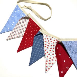 Peut inclure: Une guirlande de fanions aux couleurs patriotiques, compos&eacute;e de fanions triangulaires rouges, blancs et bleus. Les fanions pr&eacute;sentent des motifs d'&eacute;toiles et de fleurs, enfil&eacute;s sur un ruban cr&egrave;me.