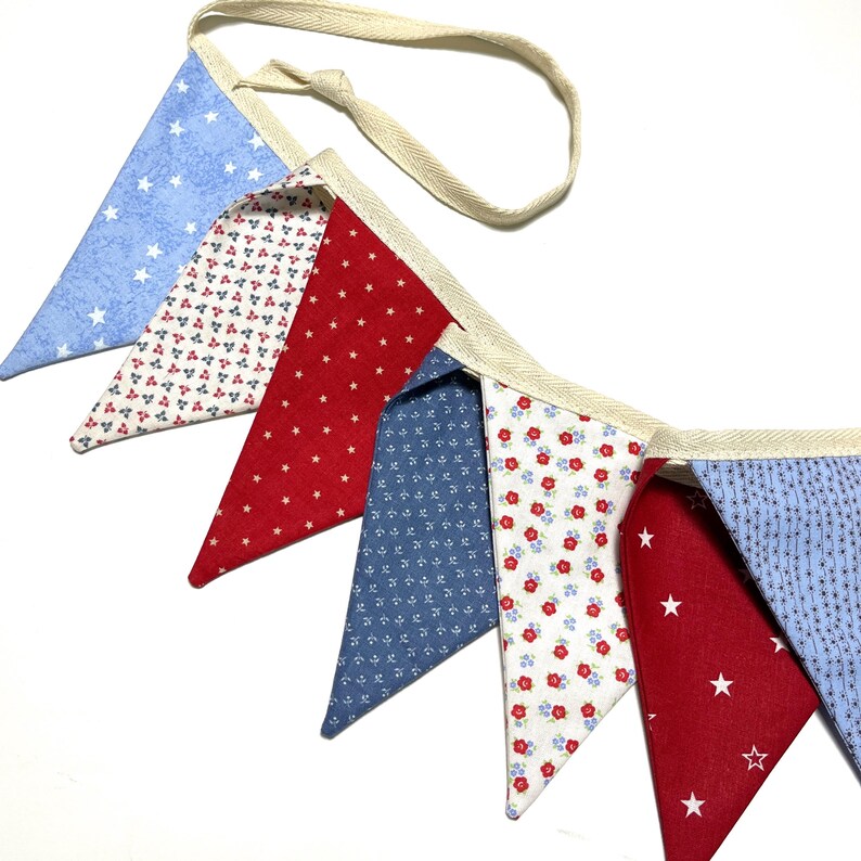Peut inclure: Une guirlande de fanions aux couleurs patriotiques, compos&eacute;e de fanions triangulaires rouges, blancs et bleus. Les fanions pr&eacute;sentent des motifs d'&eacute;toiles et de fleurs, enfil&eacute;s sur un ruban cr&egrave;me.