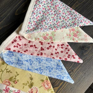 May include: A close-up of a decorative bunting with various floral patterns. The fabric triangles feature designs in pink, red, blue, and yellow, with a cream-colored trim. The bunting is made of fabric.