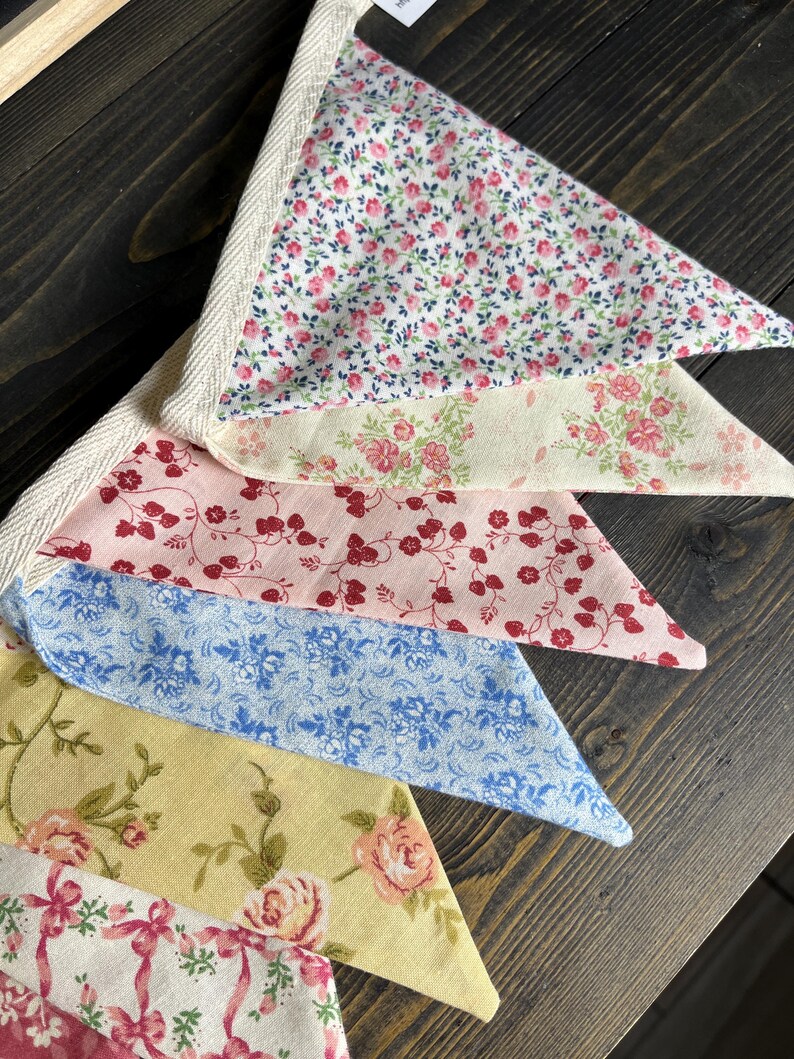 May include: A close-up of a decorative bunting with various floral patterns. The fabric triangles feature designs in pink, red, blue, and yellow, with a cream-colored trim. The bunting is made of fabric.