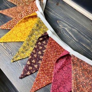 May include: A colorful bunting banner with triangular flags in shades of orange, yellow, and red. Each flag features a floral pattern. The banner is strung on a white ribbon, perfect for festive decor.