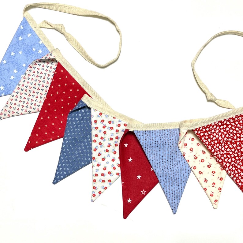 Peut inclure: Une guirlande de fanions color&eacute;s avec des drapeaux triangulaires rouges, blancs et bleus. Les fanions pr&eacute;sentent des motifs d'&eacute;toiles et de fleurs, reli&eacute;s par un ruban cr&egrave;me. Parfait pour les d&eacute;corations festives.