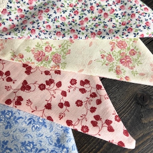 May include: Close-up of four fabric swatches with floral patterns. The fabrics feature a variety of colors, including pink, red, blue, and cream. The patterns include small flowers and vines, creating a vintage aesthetic.