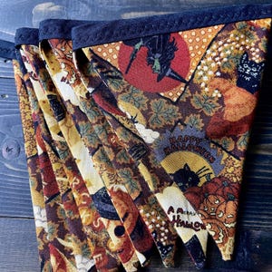 May include: A Halloween-themed bunting banner with a dark blue trim. The fabric features a repeating pattern of pumpkins, black cats, witches, and the text "Happy Halloween" and "A Merry Halloween". The color palette includes orange, brown, and black.