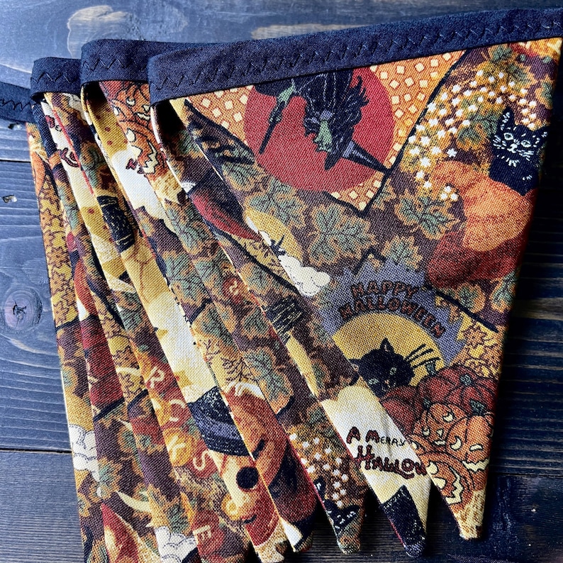May include: A Halloween-themed bunting banner with a dark blue trim. The fabric features a repeating pattern of pumpkins, black cats, witches, and the text "Happy Halloween" and "A Merry Halloween". The color palette includes orange, brown, and black.
