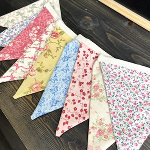 May include: A colorful fabric bunting with various floral patterns. The pennants feature designs in pink, yellow, blue, and white, with lace trim along the top edge. The bunting is arranged on a dark wooden surface.