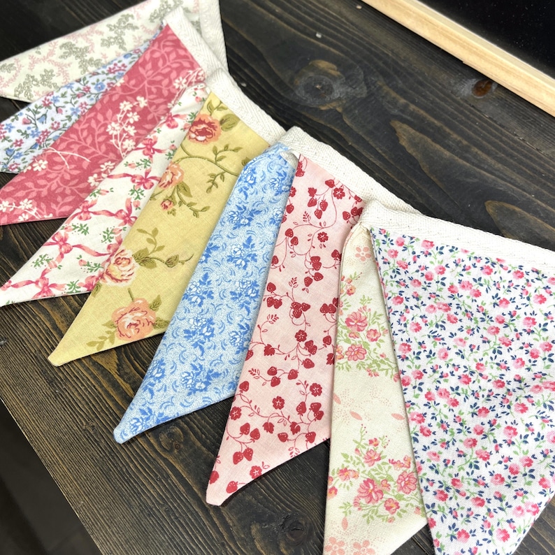 May include: A colorful fabric bunting with various floral patterns. The pennants feature designs in pink, yellow, blue, and white, with lace trim along the top edge. The bunting is arranged on a dark wooden surface.
