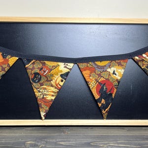 May include: A Halloween-themed pennant banner with triangular flags. The flags feature a repeating pattern of pumpkins, black cats, and other spooky imagery in orange, brown, and black. The banner is trimmed with black fabric and is displayed against a black background.