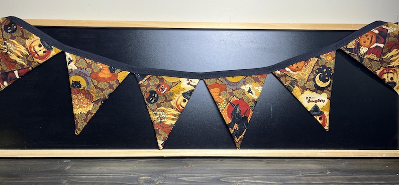 May include: A Halloween-themed pennant banner with triangular flags. The flags feature a repeating pattern of pumpkins, black cats, and other spooky imagery in orange, brown, and black. The banner is trimmed with black fabric and is displayed against a black background.