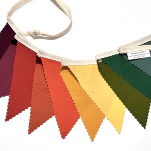 K&ouml;nnte beinhalten: Ein Stoffbanner mit zehn dreieckigen Flaggen in den Farben Burgunderrot, Orange, Gelb und Gr&uuml;n. Die Flaggen sind mit wei&szlig;em Stoff zusammengen&auml;ht und haben eine wei&szlig;e Schnur zum Aufh&auml;ngen.