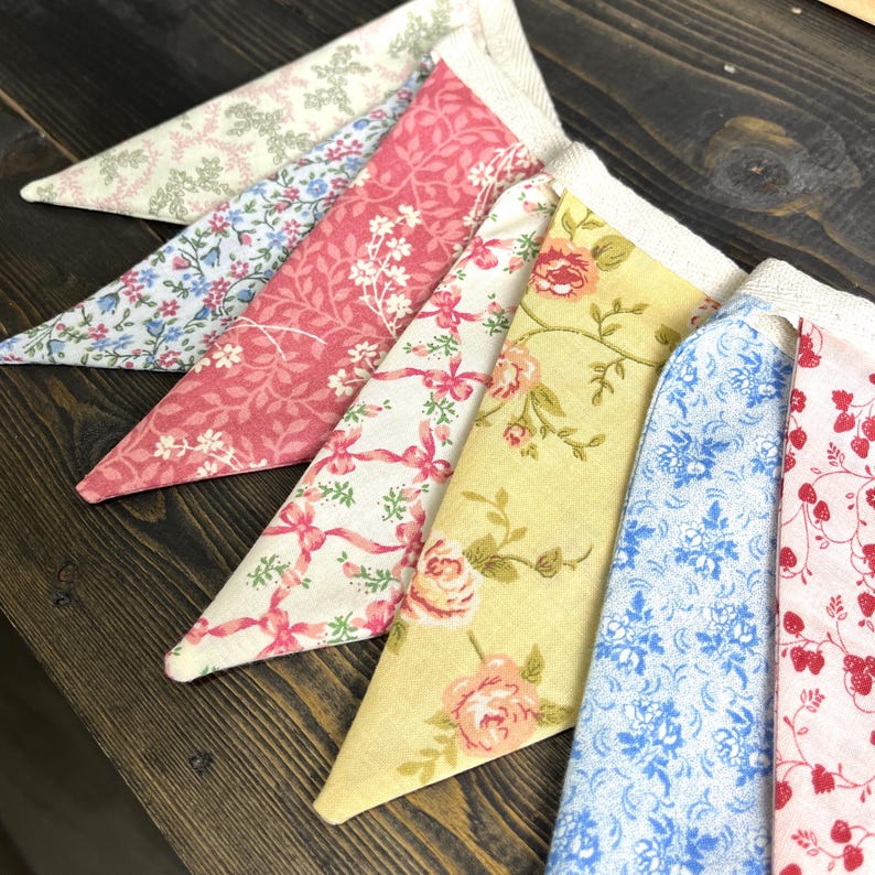 May include: A collection of colorful fabric pennants with floral and patterned designs. The pennants are in shades of pink, blue, yellow, and white, with lace trim along the top edge. The pennants are arranged in a row, creating a decorative display.