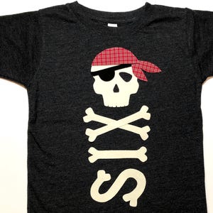 Könnte beinhalten: Ein dunkelgraues T-Shirt mit einem weißen Filz-Piratenkopf und Knochen-Design. Der Schädel trägt ein rot-weiß kariertes Bandana und eine Augenklappe. Die Zahl "sechs" ist unter dem Schädel und den Knochen geschrieben.