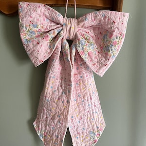 May include: A large decorative bow made from quilted fabric featuring a patchwork design. The textile displays a soft pink color palette with intricate floral patterns in shades of blue, green, and white. The bow has two long hanging tails.