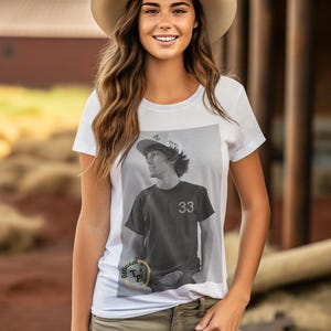 May include: White t-shirt featuring a black and white photograph of a person wearing a baseball cap and a t-shirt with the number 33 on it. The t-shirt has the text "Official T.P.I." printed on the bottom.