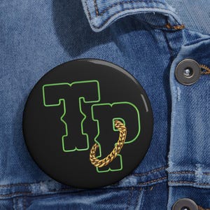 Op de afbeelding: Een zwarte knop met een groene omtrek van de letters "TP" met een gouden ketting die om de "P" is gewikkeld.