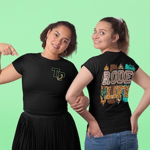 May include: Two women wearing black t-shirts. The woman on the left is wearing a t-shirt with a green and gold graphic of a scorpion and the letters "TP". The woman on the right is wearing a t-shirt with the text "Rode Life" in brown, gold, and turquoise letters with a leopard print background.