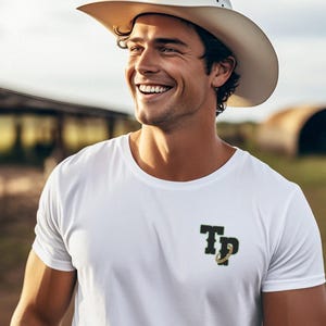 Könnte beinhalten: Ein weißes T-Shirt mit einem schwarzen und grünen Logo, auf dem "TP" mit einer goldenen Kette steht. Die Person, die das Shirt trägt, lächelt und trägt einen weißen Cowboyhut.