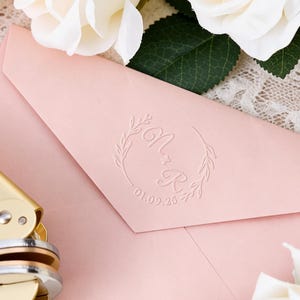 May include: A gold-colored wax seal stamp and a light pink envelope with a monogram and date. The envelope is surrounded by white and pink roses and rests on a lace surface. The seal stamp is next to the envelope.