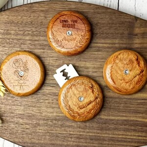 May include: Four wooden razor blade holders with engraved designs and text, including "LOVE YOU MOM" and floral patterns. The holders are circular, with a small metal center, and rest on a dark wooden cutting board.
