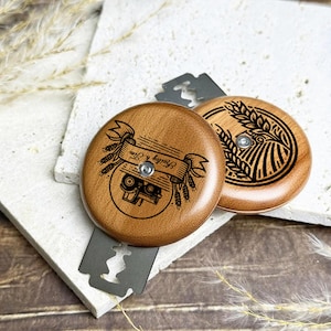 Könnte beinhalten: Zwei runde Rasierklingenhalter aus Holz mit eingravierten Designs. Einer zeigt ein Banner mit Text und einem Traktor, der andere ein Weizenfeld. Die Halter sind braun und liegen auf einer weißen Oberfläche.