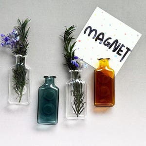 May include: Four glass bottles of varying colours and designs are arranged on a grey surface. Two bottles contain greenery and flowers, one is teal, and the last is amber. A card with the word "MAGNET" is also present.