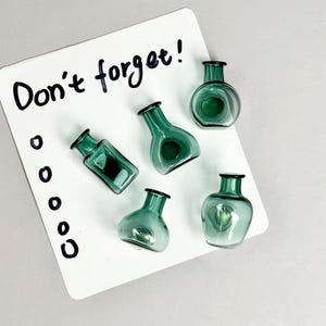 Puede incluir: Vista cenital de seis pequeños jarrones de cristal de color verde azulado con bordes negros. Están colocados sobre un bloc de notas blanco con las palabras "¡No te olvides!" escritas con tinta negra. El bloc de notas también tiene una serie de círculos negros.