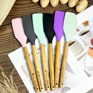 Może przedstawiać: Sześć kolorowych silikonowych szpatułek z drewnianymi uchwytami. Główki szpatułek są różowe, szare, miętowo-zielone, czarne, fioletowe i jasnozielone. Każdy uchwyt ma inny wygrawerowany napis, np. "Mom's Kitchen" i "World's Best Cook".