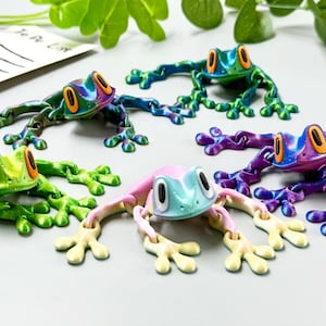 May include: A collection of colorful frog figurines in various shades of green, blue, purple, and pink. Each frog has large, orange eyes and is posed with its legs outstretched. The frogs are arranged on a light gray surface.