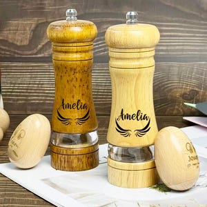 May include: Two wooden salt and pepper grinders with clear glass bases. The grinders are engraved with the name "Amelia" and a wing design. Two wooden egg-shaped objects are also visible.