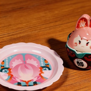 Puede incluir: Un plato rosa con borde festoneado y un recipiente decorativo con diseño de conejo. El plato presenta un gráfico rosa y azul. El recipiente es rosa con base negra y la palabra "moon" en inglés y japonés.