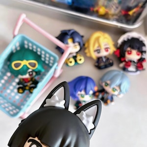 Puede incluir: Una colección de figuras de estilo anime con varios colores de cabello y atuendos. Una figura está en una pequeña cesta de la compra azul. Las figuras tienen rasgos faciales detallados y están dispuestas sobre una superficie blanca.