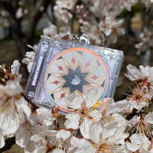 Könnte beinhalten: Ein transparentes CD-Gehäuse aus Kunststoff mit einem silbernen Schlüsselanhänger. Die CD zeigt einen schwarzen Stern mit goldenen und roten Akzenten auf weißem Hintergrund. Der Text "BARDICNOEG" ist auf der Seite des Gehäuses aufgedruckt.