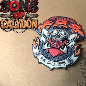 May include: A metal pin featuring a stylized boar wearing goggles, with the text "SONS OF CALYDON" and Japanese characters. The pin has red, orange, silver, and black colours.