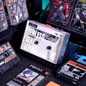 May include: A clear cassette player with the text "ZENLESS ZONE ZERO" is displayed, surrounded by cassette tapes and promotional cards. The tapes and cards feature anime-style illustrations and the game's logo. The color scheme includes black, orange, and white.