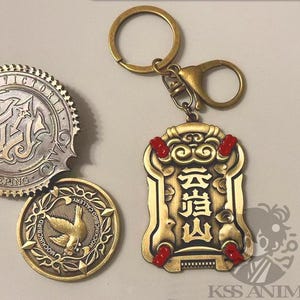 Puede incluir: Un llavero dorado con un colgante rectangular con caracteres chinos y detalles rojos. También se ven dos monedas doradas con diseños intrincados, una con un pájaro y la otra con texto.