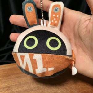 May include: A plush bunny keyring with a round head and orange accents. The bunny has large green eyes, a black face, and orange ears with blue and white details. The keyring has the text "W1." on the front.