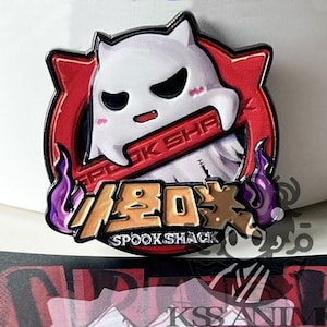 Puede incluir: Un pin redondo de esmalte con un personaje fantasma blanco con ojos negros y boca roja, sobre un fondo rojo. El pin incluye el texto "SPOOK SHACK" y caracteres japoneses. Llamas moradas acentúan el diseño.