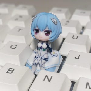 May include: A small figurine of a character with blue hair and a white and blue outfit, resting on a white surface. The figurine has large purple eyes and is set against a backdrop of white keyboard keys.