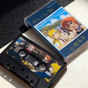Puede incluir: Una cinta de cassette negra con ilustraciones coloridas de estilo anime y texto japonés, junto con una funda de plástico transparente con obras de arte similares. La funda presenta el texto "We will Kessoku Band" y otros caracteres japoneses.