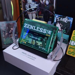 May include: A green cassette tape-shaped audio player with the word "ZENLESS" printed on it, resting on a white box. The player has clear plastic headphones attached. The box reads "KMIDO Audio Studio CASSETTE @PLAYER".
