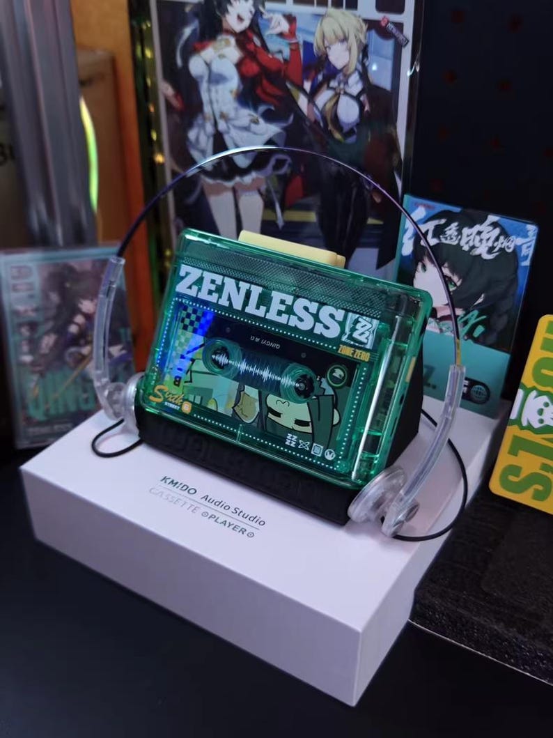 May include: A green cassette tape-shaped audio player with the word "ZENLESS" printed on it, resting on a white box. The player has clear plastic headphones attached. The box reads "KMIDO Audio Studio CASSETTE @PLAYER".