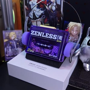 May include: A purple cassette tape player with the text "ZENLESS" and "ZONE ZERO" on the front. The player has purple ear pads and is sitting on a white box. The box has the text "KMIDO Audio Studio CASSETTE @PLAYER".