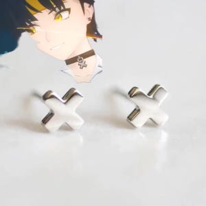 Puede incluir: Un par de pendientes de botón plateados con forma de X. Los pendientes son pequeños y tienen un acabado pulido. El fondo es blanco neutro y suave.